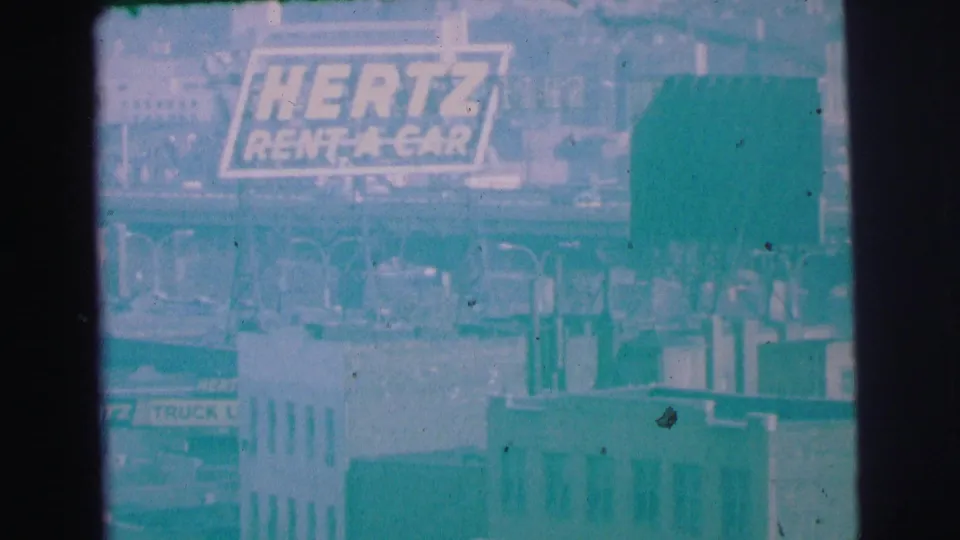 1973 Lynbrook, New York frame with the Hertz Rent A Car sign clearer above road barriers, vehicles, and low commercial buildings.