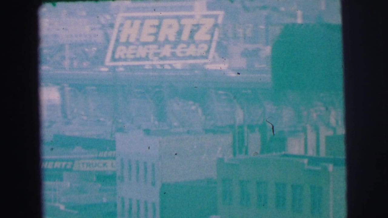 1973 Lynbrook, New York 8mm frame with a visible Hertz Rent A Car sign, soft building facades, and dark film-frame edges.
