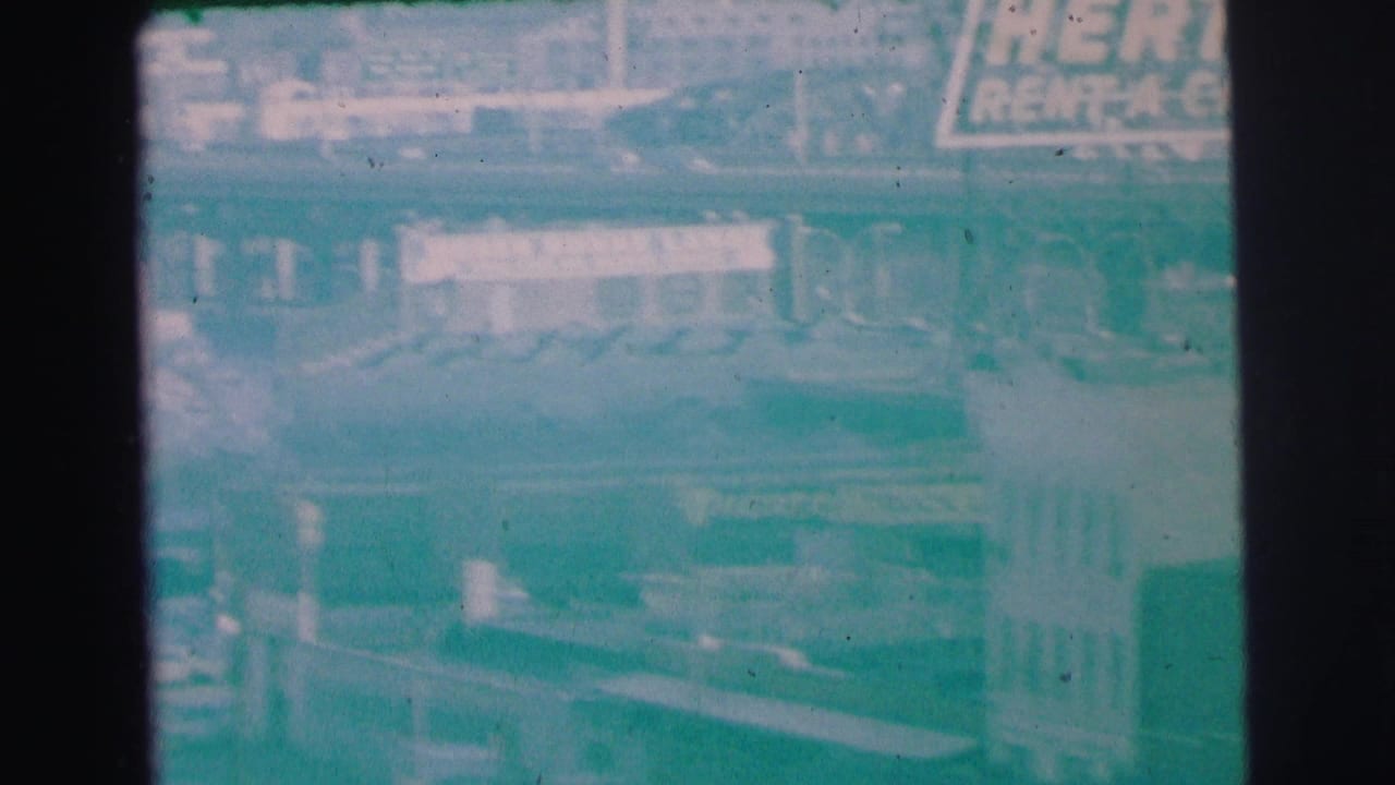 1973 Lynbrook 8mm view from above, showing Hertz signage near storefronts and a horizontal bridge or roadway edge.