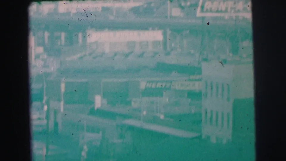 1973 Lynbrook home-movie frame showing Hertz Rent A Car signage among buildings, with visible film grain and frame borders.