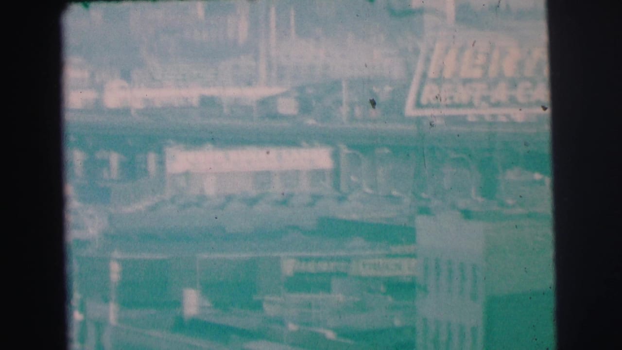 Soft 8mm view of a Hertz Rent-A-Car sign above a hazy commercial district.