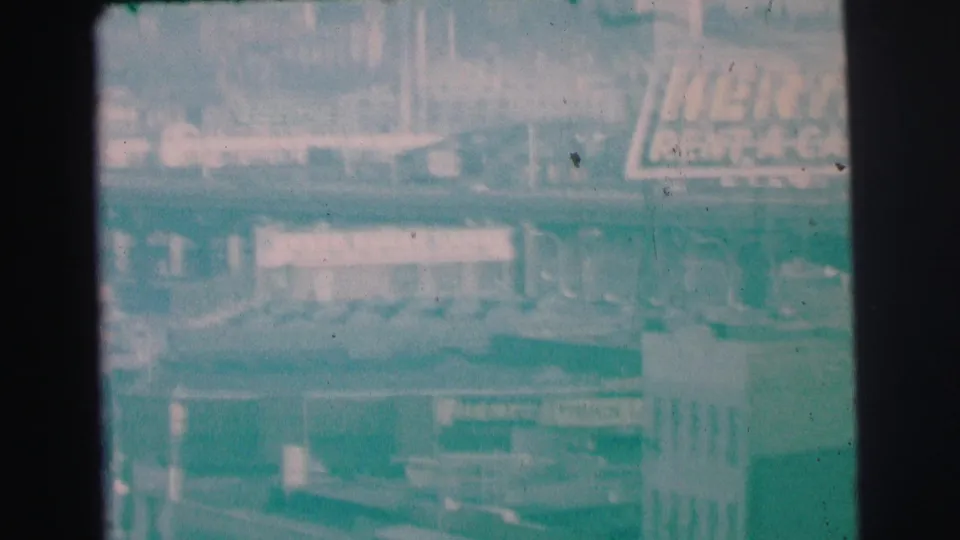 1973 Lynbrook home-movie frame showing a large Hertz Rent-A-Car sign, low buildings, and parked vehicles through haze.