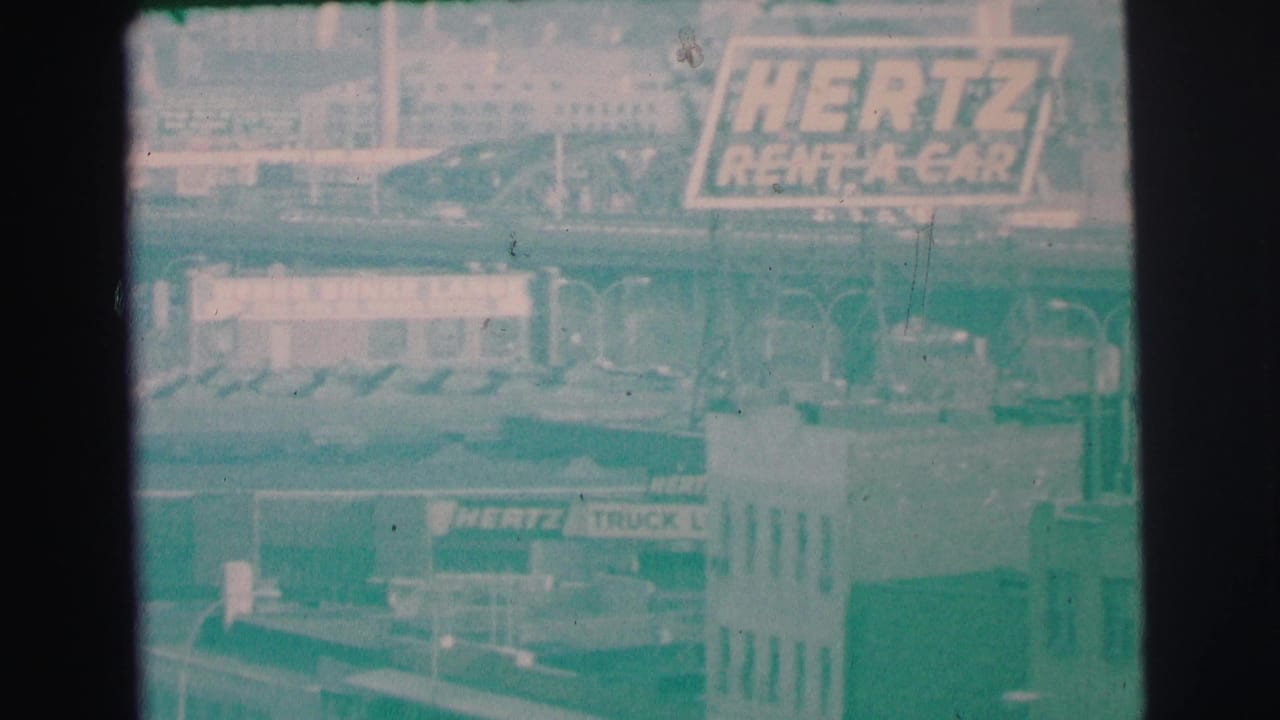 1973 Lynbrook frame with Hertz Rent-A-Car signage, a truck rental sign, low buildings, and distant rooftops in haze.