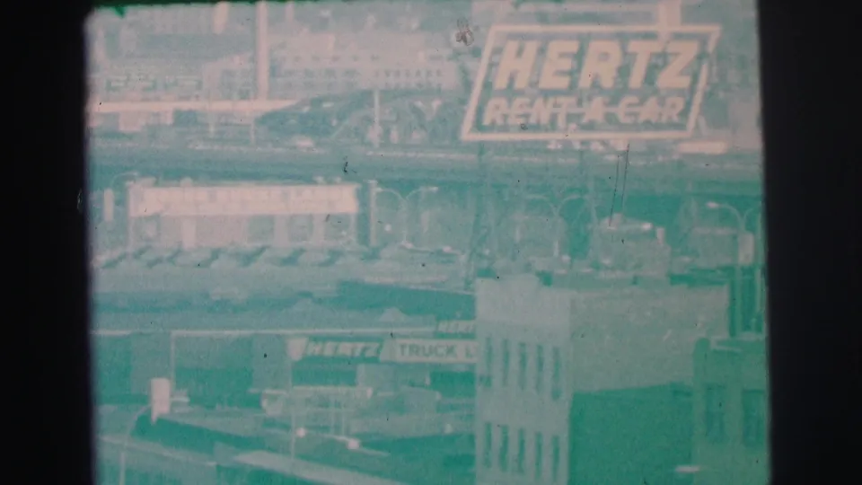 1973 Lynbrook frame with Hertz Rent-A-Car signage, a truck rental sign, low buildings, and distant rooftops in haze.