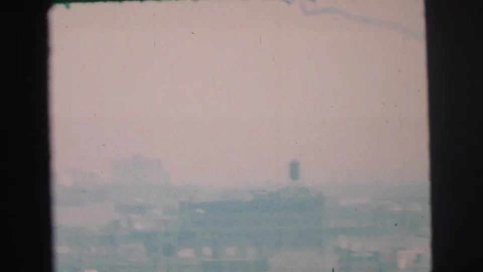 1973 Lynbrook home-movie frame with a washed, hazy view of low buildings and a distant water tower-like shape.