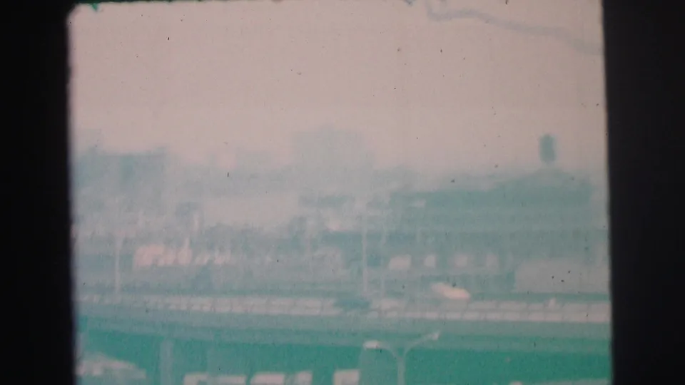 1973 Lynbrook, New York home-movie frame showing a blurred roadway scene with low buildings, a distant tower form, and faded teal 8mm color.