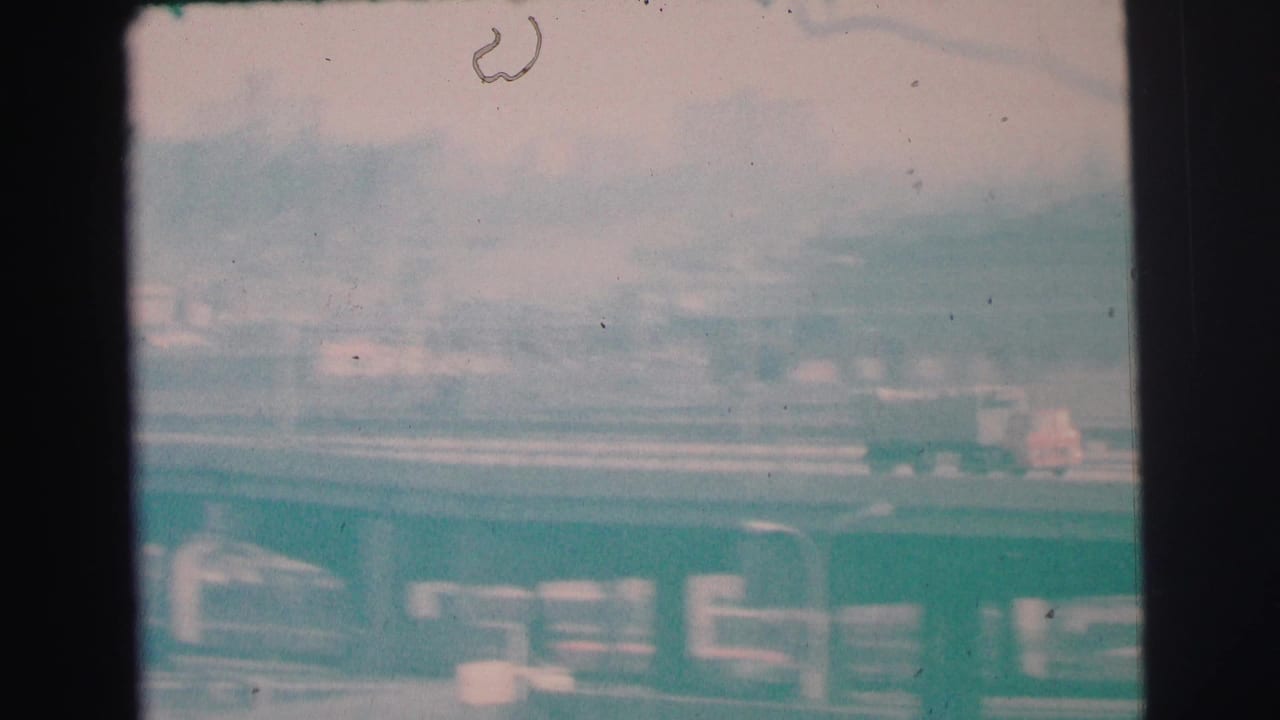 Faded 1973 Lynbrook frame with cars and a truck on a roadway, low buildings beyond, and visible 8mm dust and frame edges.