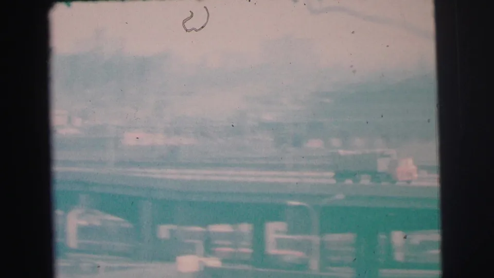 Faded 1973 Lynbrook frame with cars and a truck on a roadway, low buildings beyond, and visible 8mm dust and frame edges.