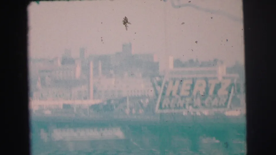 1973 Lynbrook home-movie frame showing a Hertz Rent-A-Car billboard near traffic, with buildings behind and heavy 8mm fading.
