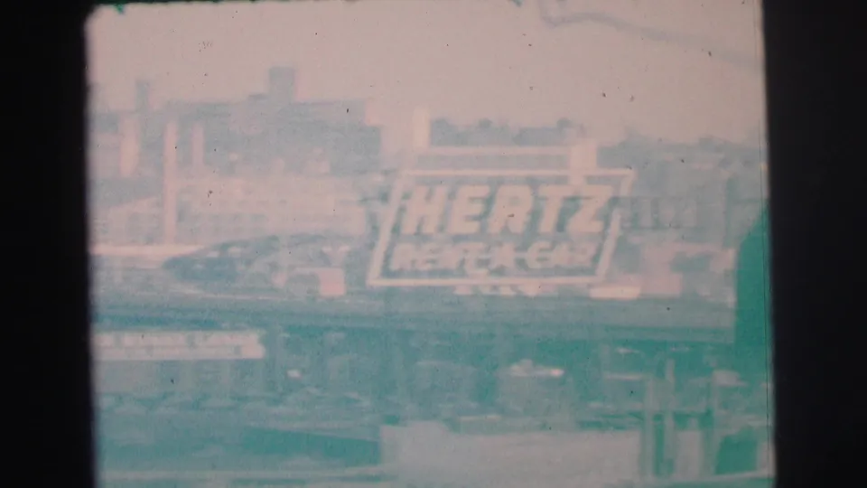 Soft 1973 Lynbrook frame with a Hertz Rent-A-Car billboard, passing vehicles, layered roadway structures, and faded teal 8mm color.