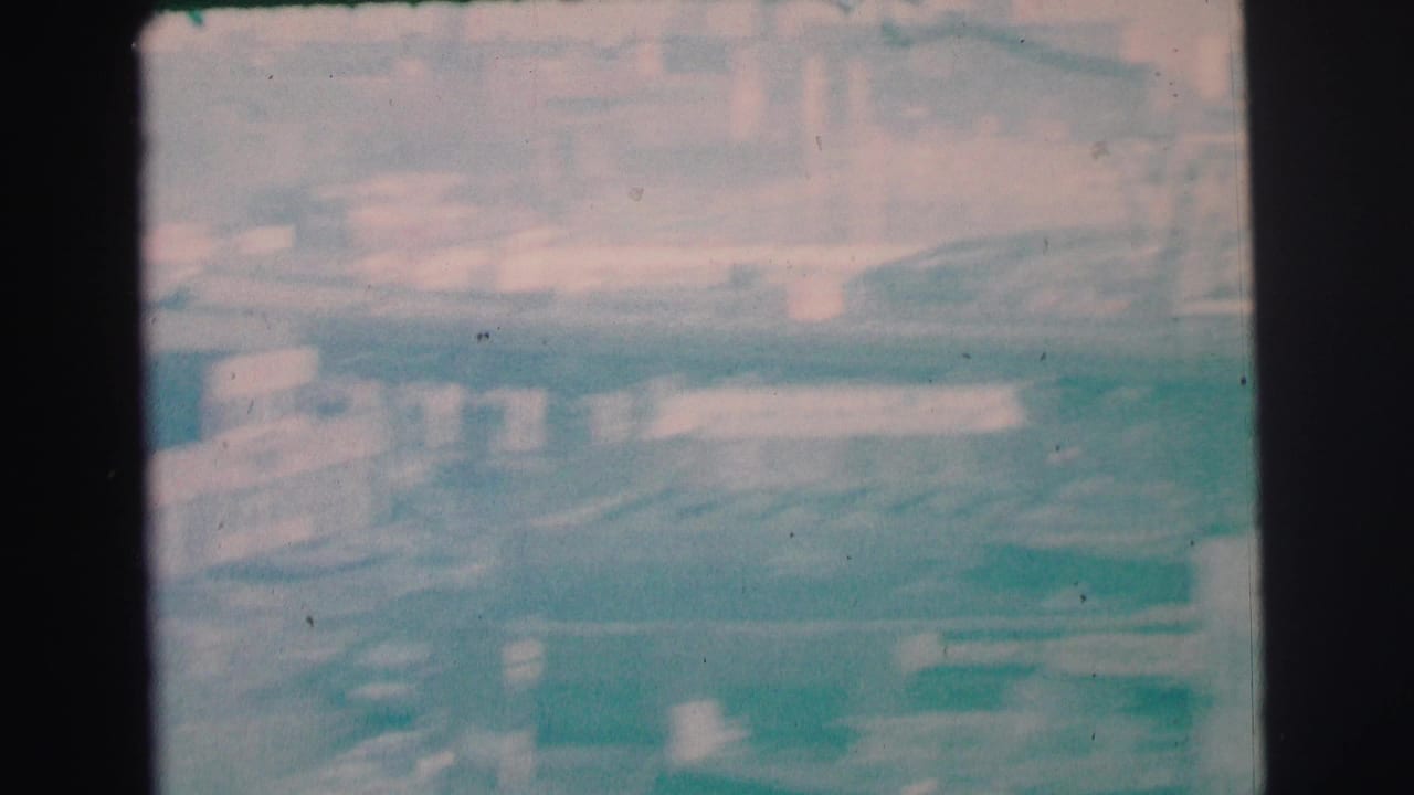 Hazy 1973 Lynbrook, New York 8mm frame showing an indistinct urban street view with buildings and parked vehicles.