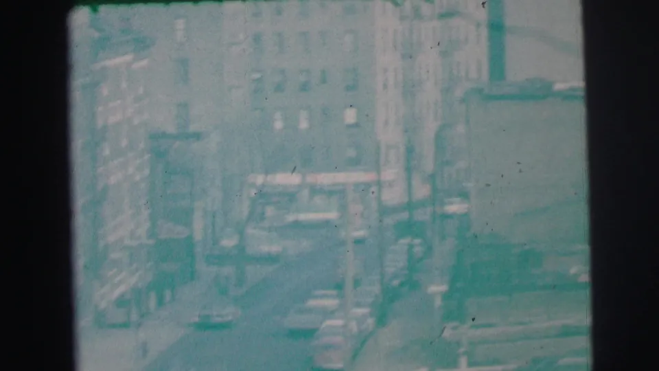 Hazy 1973 Lynbrook, New York 8mm frame showing a street between tall buildings, parked cars, and indistinct signs.