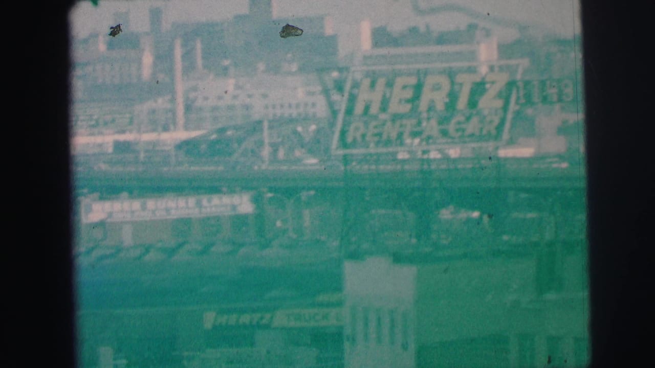 1973 Lynbrook, New York home-movie frame showing a large Hertz Rent-A-Car sign, roadway structure, and hazy urban buildings.