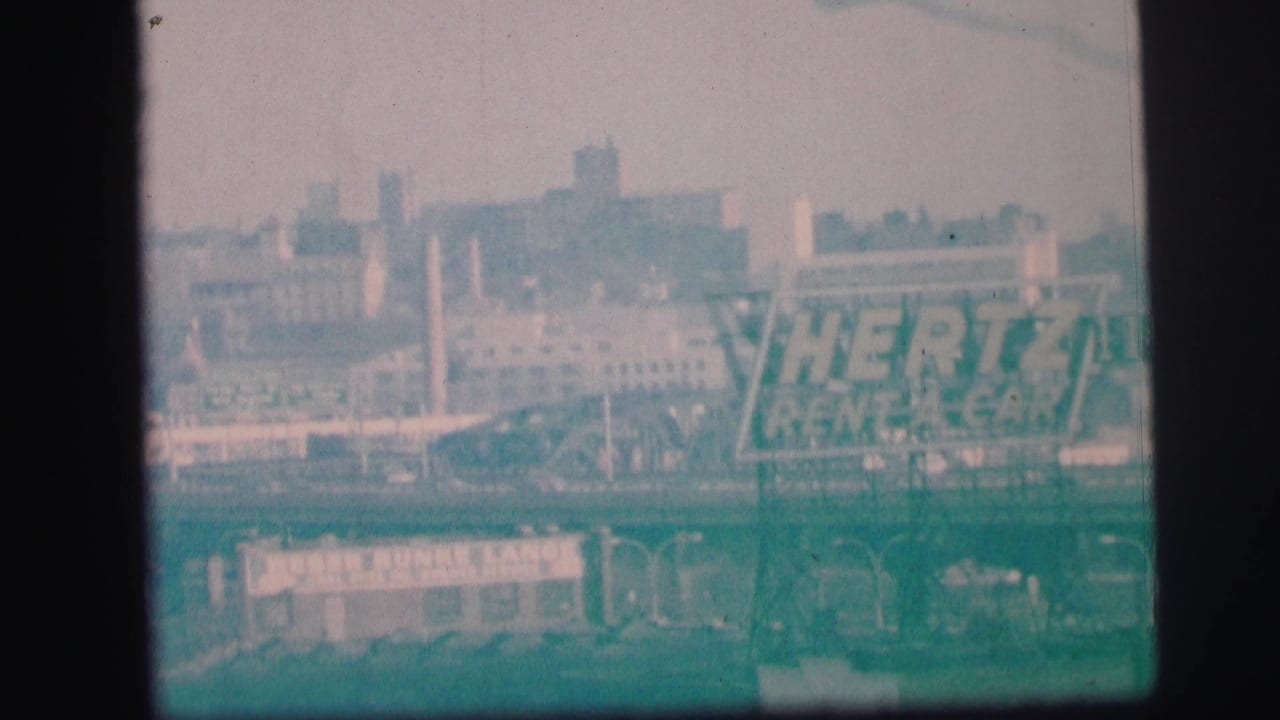 Hazy 1973 Lynbrook frame with the Hertz Rent-A-Car sign in the midground, backed by commercial buildings and road structures.