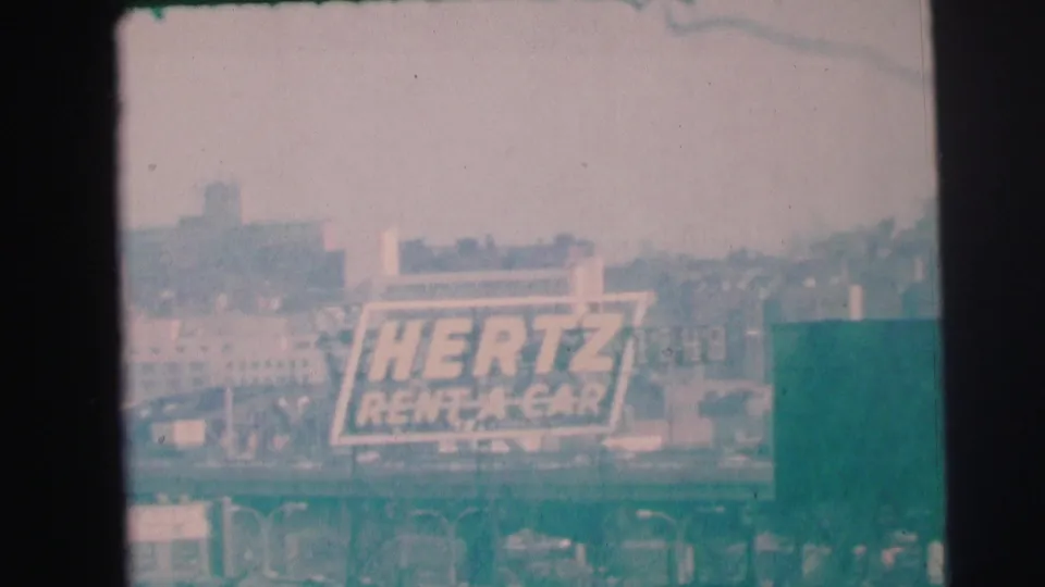 Final 1973 Lynbrook frame with the Hertz Rent-A-Car sign centered near road structures and indistinct buildings under a pale sky.