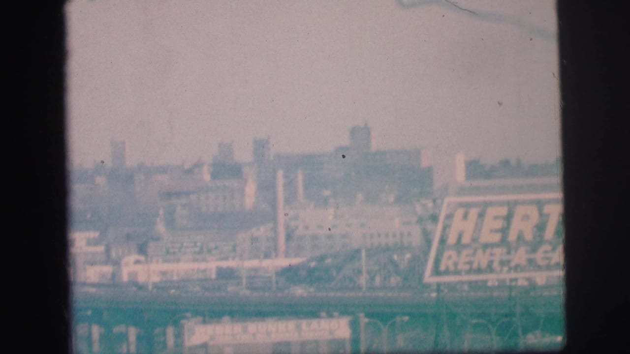 A faded 8mm view of the Hertz Rent-A-Car sign beside low industrial buildings in Lynbrook, New York.