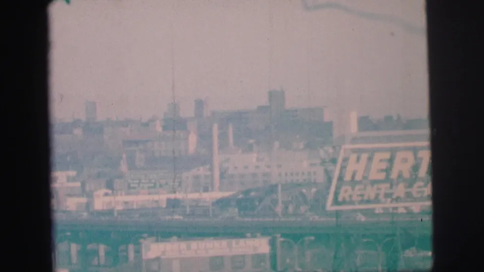 1973 8mm frame with a partial Hertz Rent-A-Car sign, hazy buildings, and a lower rail or roadway structure.