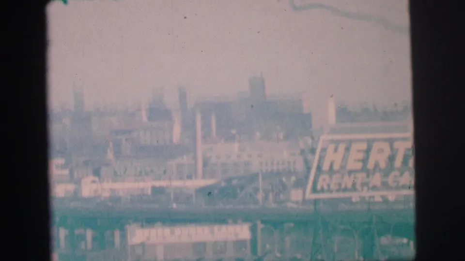 Faded 1973 Lynbrook 8mm frame showing a partial Hertz sign and distant industrial buildings under a pale sky.