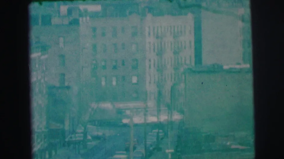 Elevated 1973 Lynbrook view showing apartment facades, fire escapes, parked or moving cars, and a city street below.