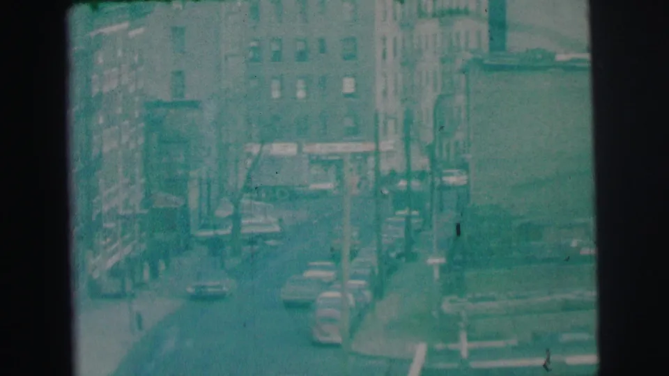 Final 1973 Lynbrook frame with multi-story buildings, traffic, utility poles, and a bus or large vehicle visible in the street scene.