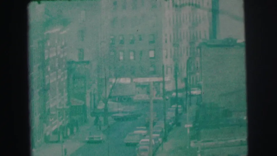 1973 Lynbrook street scene seen from above, with mid-rise buildings, cars, utility poles, and aged 8mm color shift.