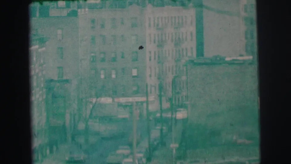 Elevated 1973 Lynbrook view showing a street corridor, older cars, apartment facades, poles, and aged 8mm texture.