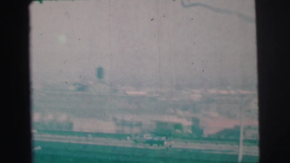 1973 Lynbrook, New York frame showing distant roads, rooftops, poles, and a small tower-like form through haze.