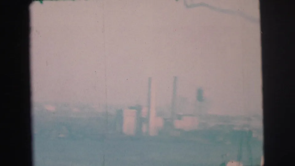 Hazy 1973 8mm view of a far industrial shoreline near Lynbrook, with smokestacks and pale bay water in soft focus.