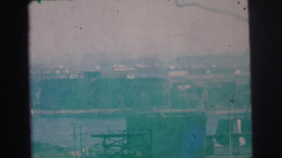 1973 Lynbrook, New York 8mm frame showing a distant shoreline, low buildings, utility forms, and water under a washed sky.