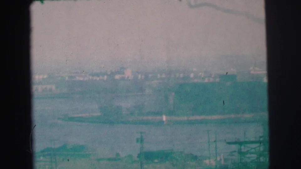 1973 Lynbrook, New York 8mm frame showing water, distant buildings, shoreline forms, and utility poles in a soft home-movie panorama.