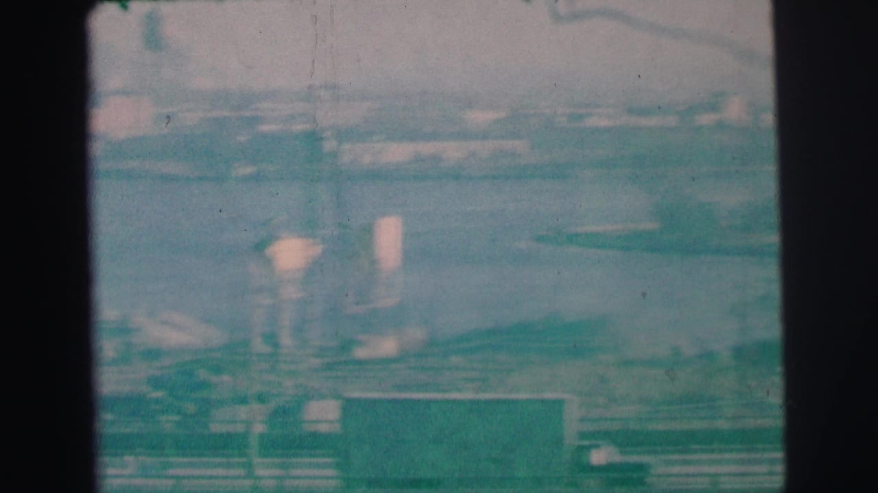 1973 Lynbrook harbor frame with water, low industrial shoreline buildings, vertical stacks or poles, and teal-tinted 8mm texture.