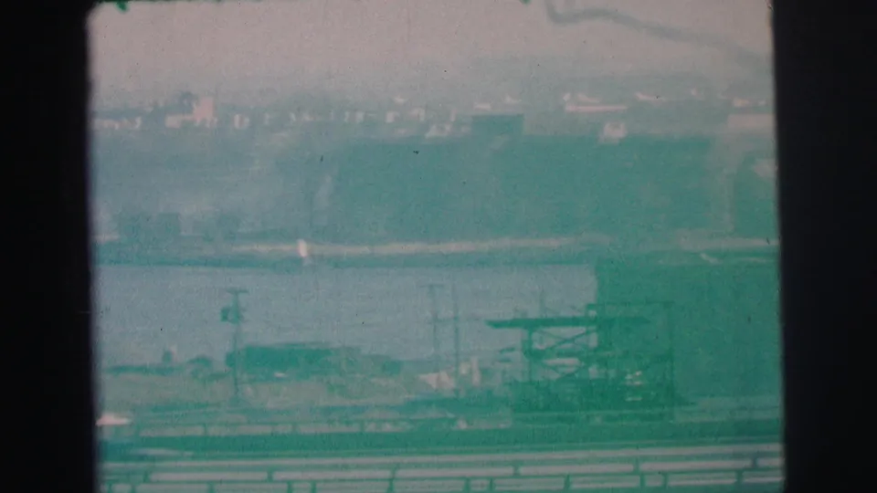 A 1973 8mm Lynbrook waterfront frame shows a small boat on the water below a hazy industrial shoreline.