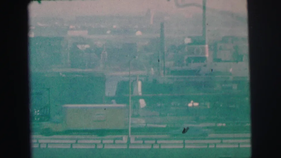 1973 Lynbrook harbor frame showing water, low waterfront structures, vertical poles or stacks, and the soft teal cast of 8mm film.