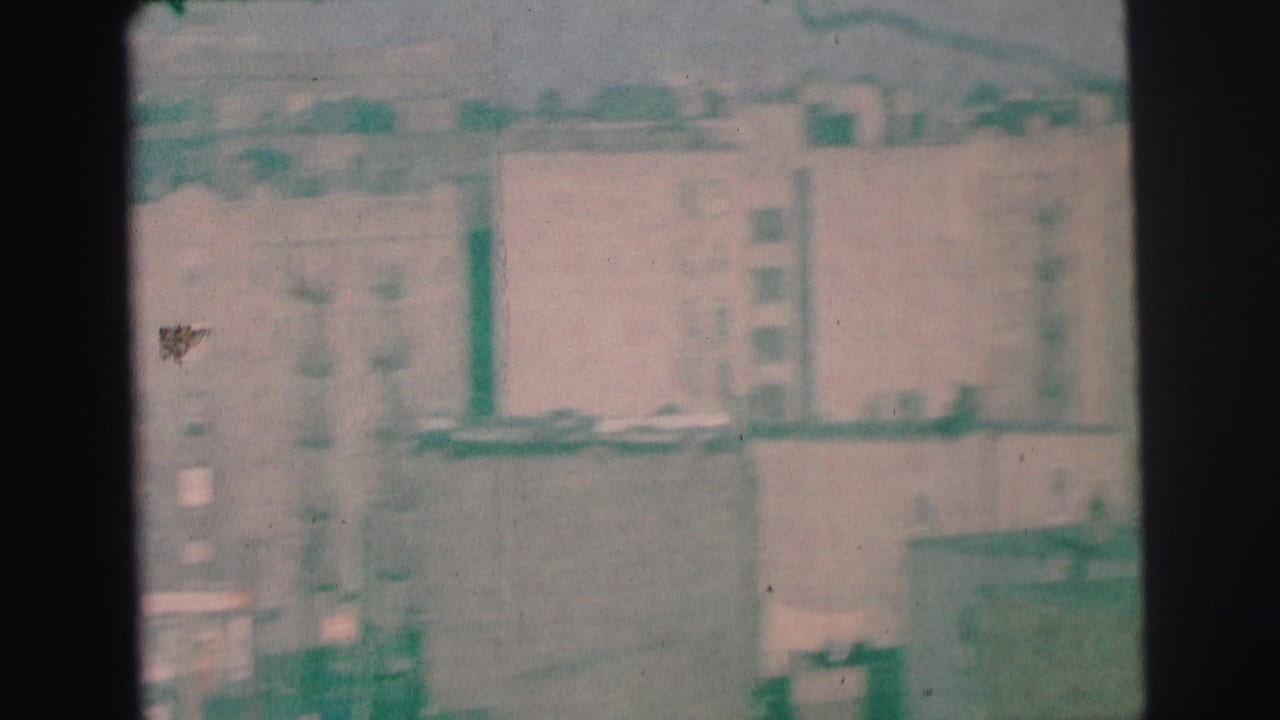 Grainy 1973 Lynbrook frame of mid-rise apartment facades, fire escapes, rooftops, and nearby street details.