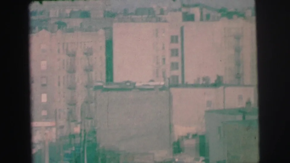 1973 Lynbrook home-movie frame showing apartment blocks, fire escapes, rooftops, and muted street details in soft focus.