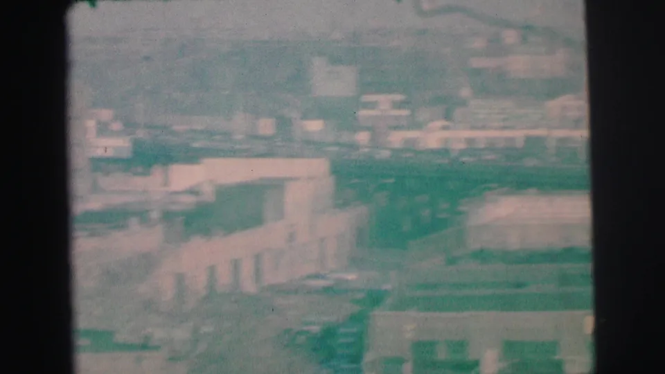 1973 Lynbrook, New York frame showing hazy rooftops, low commercial buildings, parked cars, and distant structures.