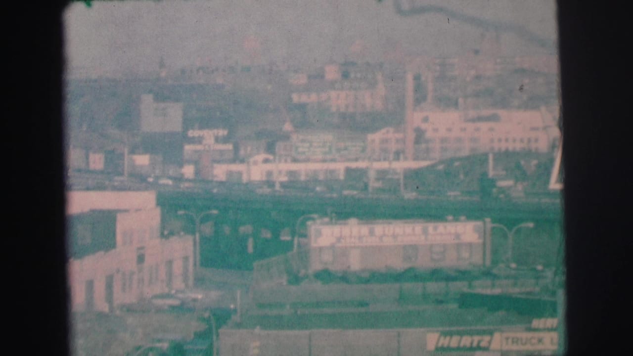 1973 Lynbrook, New York 8mm frame showing a hazy elevated view of buildings, roads, cars, and a distant urban horizon.