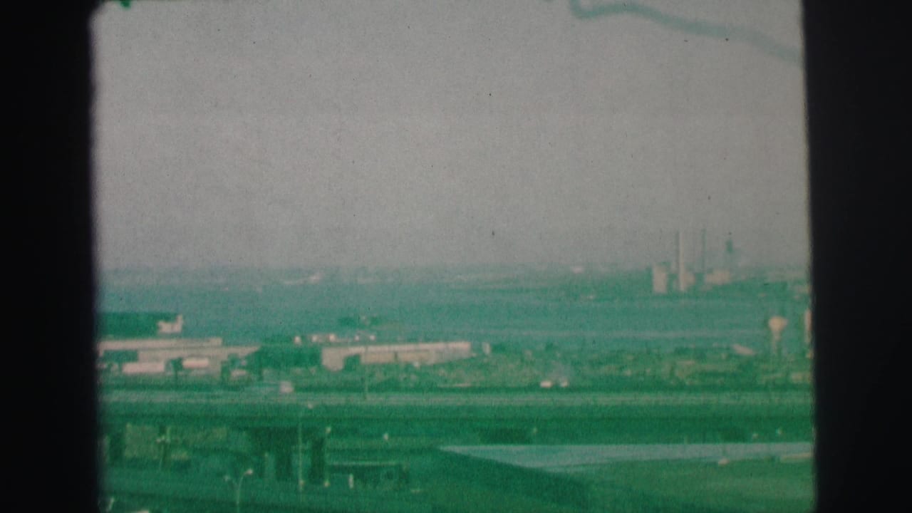 Hazy elevated view of roadway, distant buildings, smokestacks, and dark film borders.