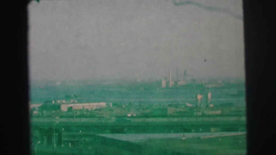 1973 Lynbrook, New York frame with a distant roadway, low buildings, and smokestacks visible through a hazy green-tinted film scan.