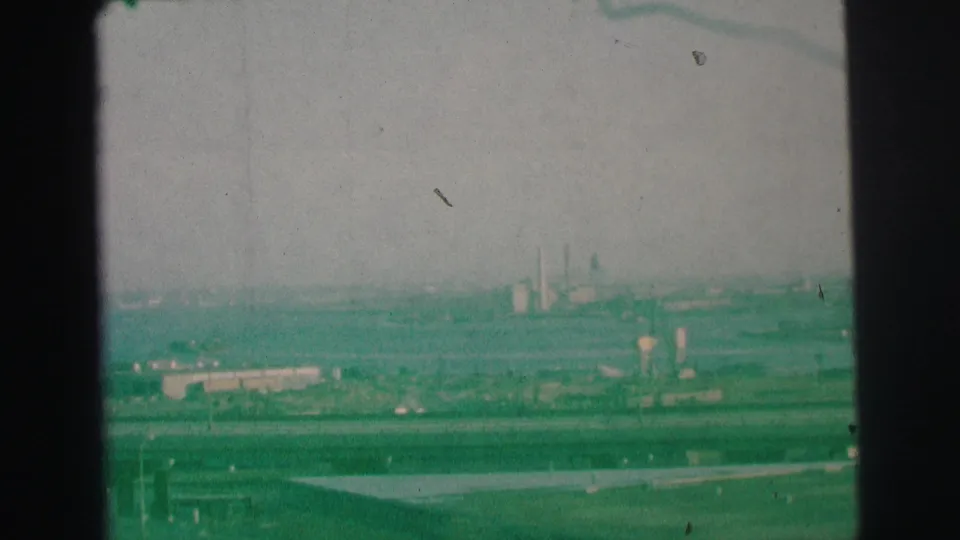 Final 1973 Lynbrook frame showing an elevated roadway view with distant industrial buildings, smokestacks, and visible 8mm dust marks.