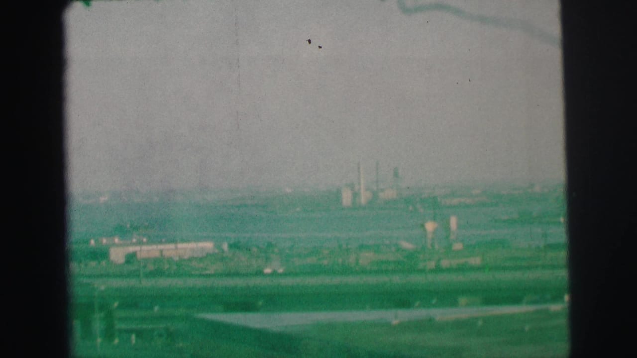 1973 Lynbrook-area home-movie frame showing a green-tinted elevated view over highways, cars, low buildings, and distant smokestacks.