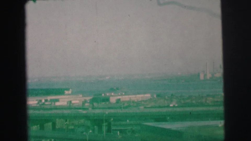 1973 Lynbrook-area 8mm frame looking over highway bridges and small cars toward low industrial structures in heavy haze.