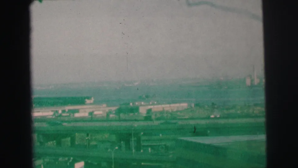 Faded 1973 Lynbrook-area frame showing motorway traffic, bridgework, low industrial buildings, and smokestacks under a hazy sky.