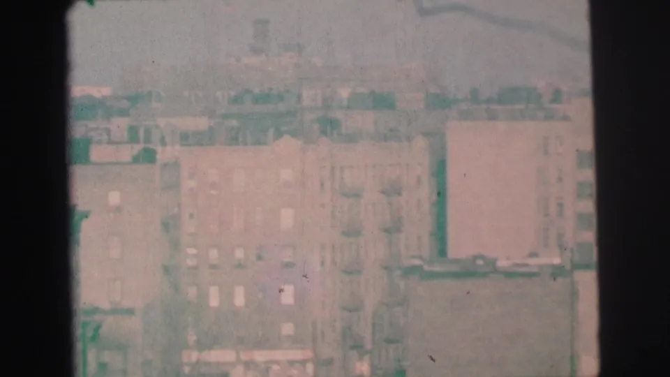 1973 Lynbrook home-movie view of hazy apartment buildings and rooftops seen through a dark vertical window frame.