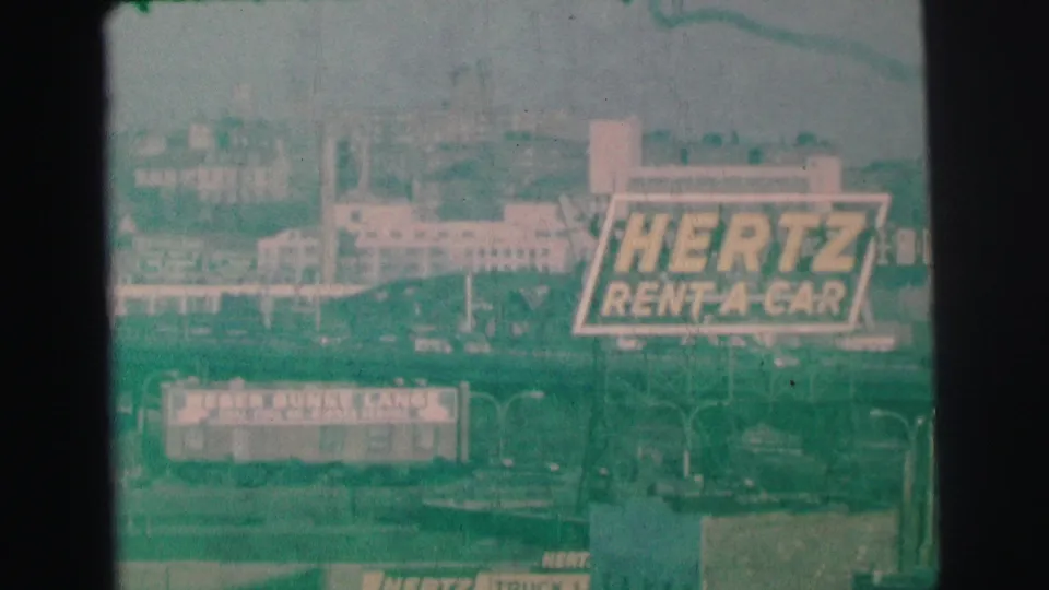 1973 Lynbrook-area frame with a large Hertz Rent-A-Car billboard, roadside structures, utility poles, and distant buildings.