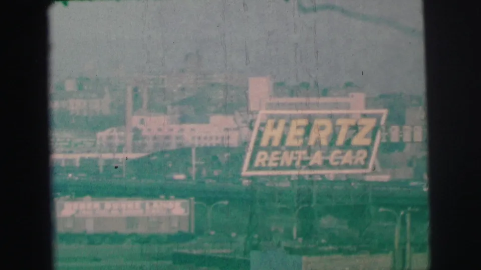Green-tinted 8mm frame of a prominent Hertz Rent-A-Car roadside billboard in Lynbrook, New York, with surrounding buildings and poles.