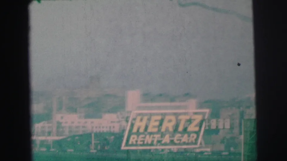 Final 8mm frame showing the Hertz sign in Lynbrook against muted buildings and a faded sky.