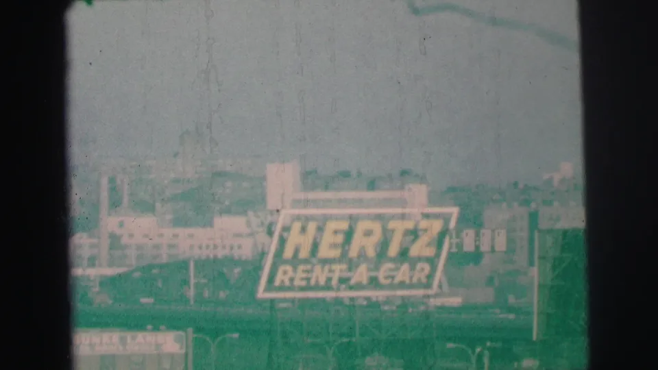 1973 Lynbrook, New York 8mm frame showing a large Hertz Rent-A-Car billboard among commercial buildings and streetlights.