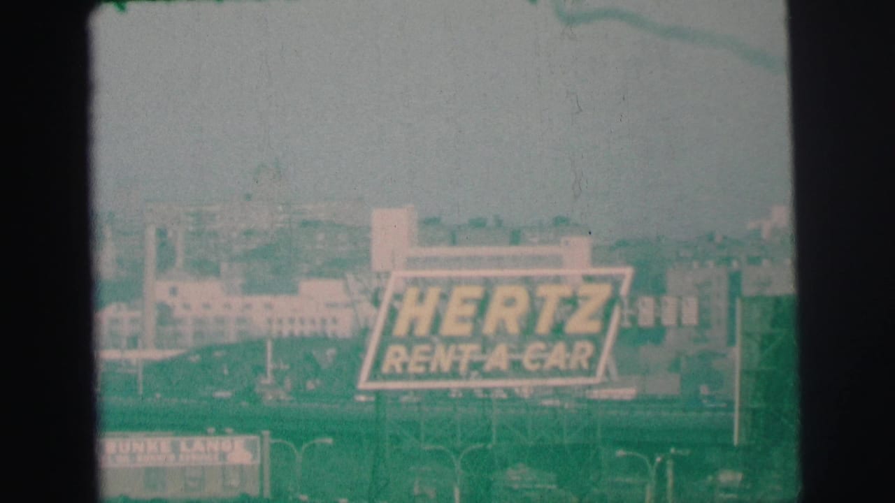 Hertz Rent-A-Car billboard centered beyond streetlights and low buildings.