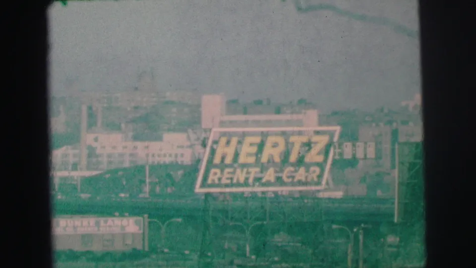 Green-tinted 1973 Lynbrook 8mm frame with a Hertz Rent-A-Car billboard above roadside signs and commercial structures.